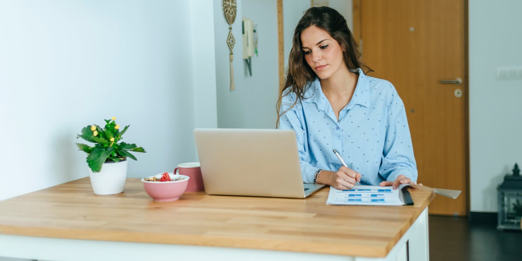 person sitting at kitchen table on laptop while writing something on paper
