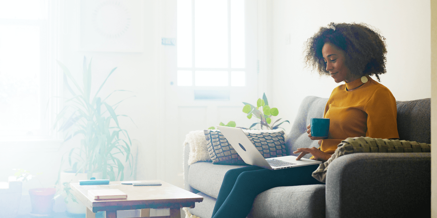 person sitting on their couch working on a laptop and drinking from a mug