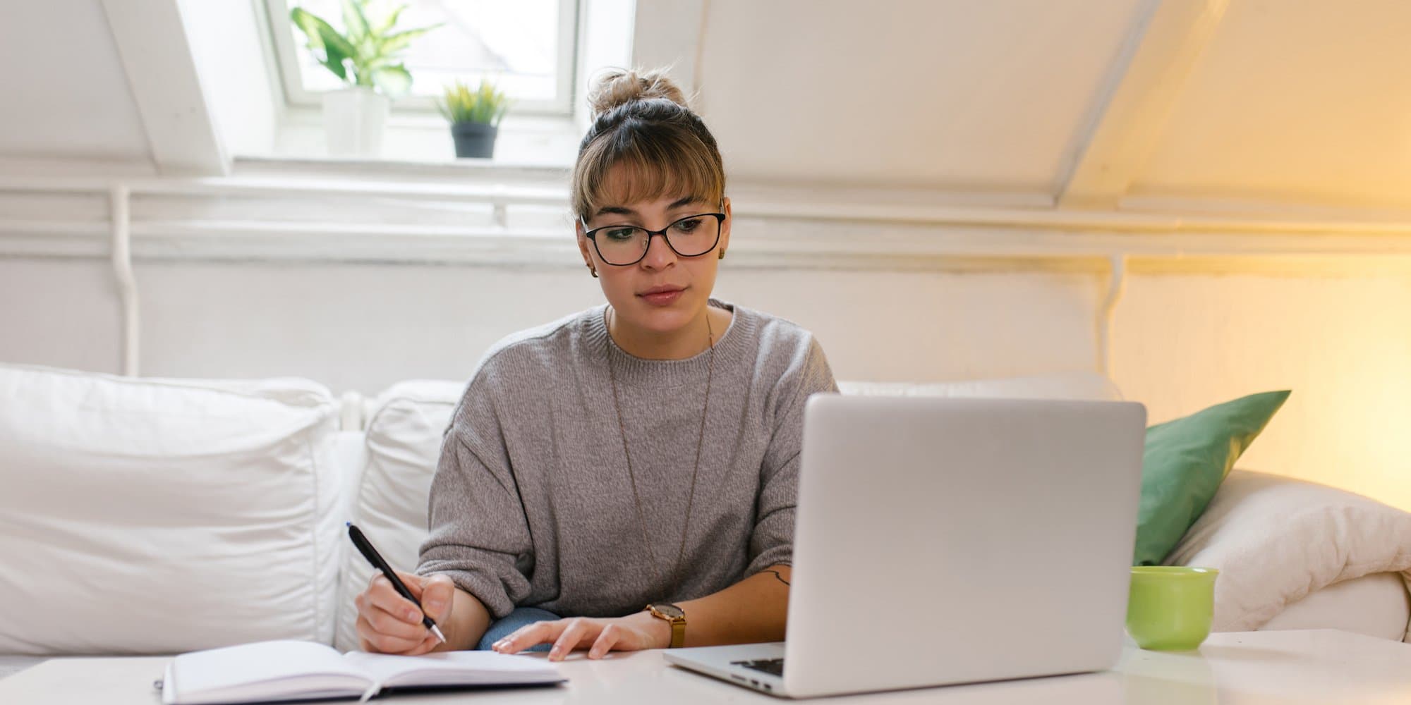 person sitting on a couch, looking at their laptop on a coffee table while writing in a notebook