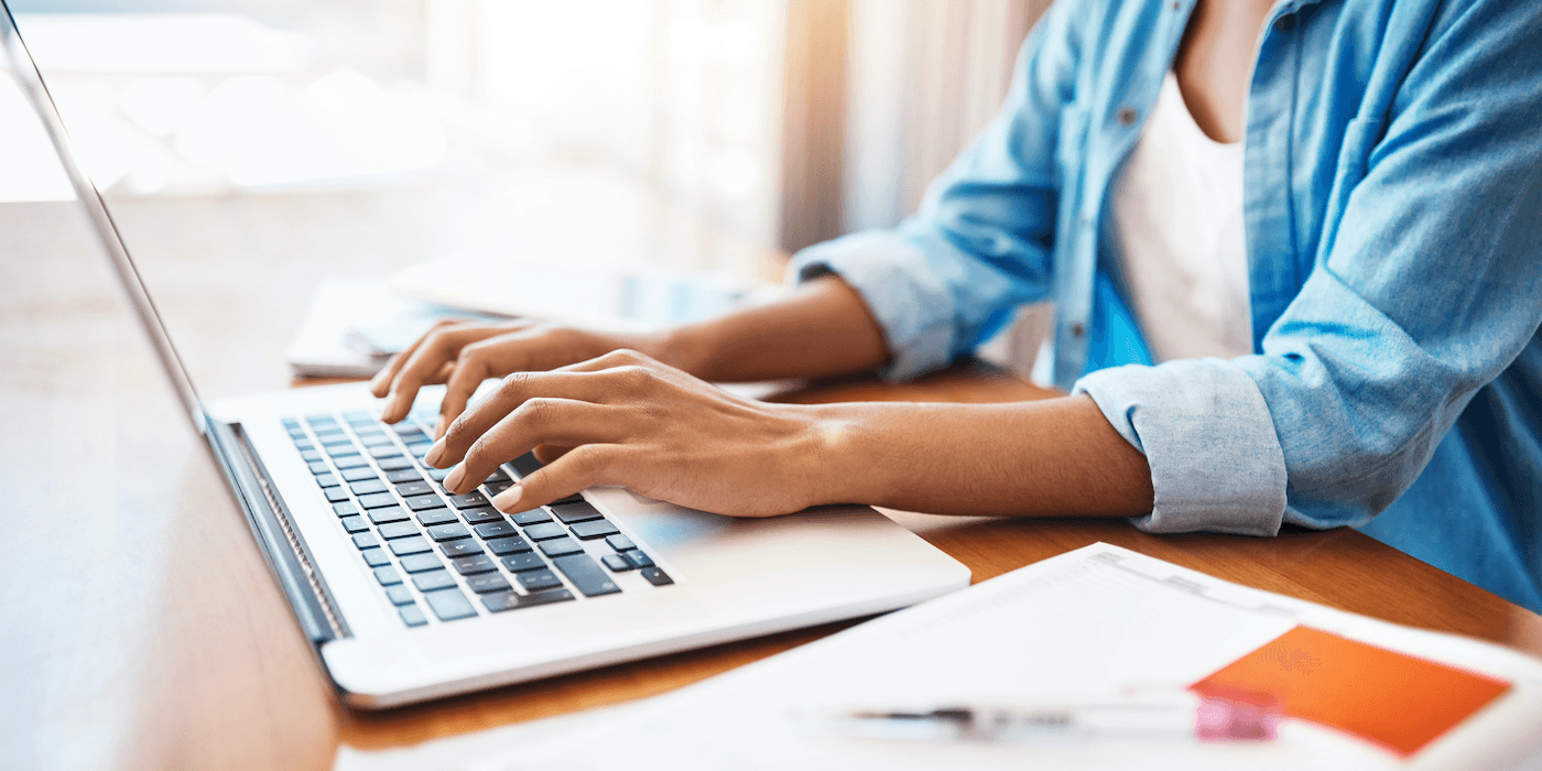 hands seen typing on a laptop with a notebook open nearby