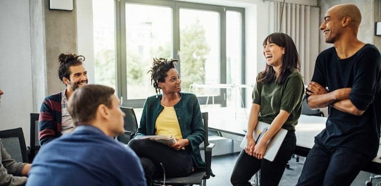 people sitting to the side of a conference table, some in chairs, some sitting on tables, talking and smiling