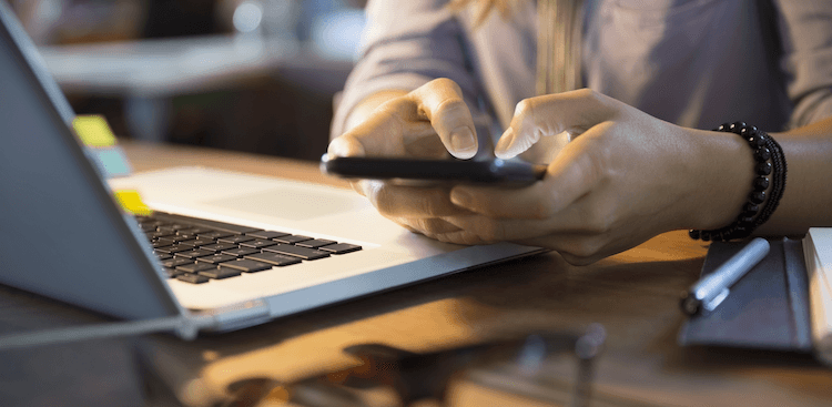 person using phone with laptop open and a pile of papers and glasses on the desk