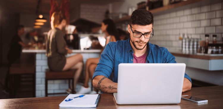 person sitting at a coffee shop working on a laptop with a notebook and cell phone nearby