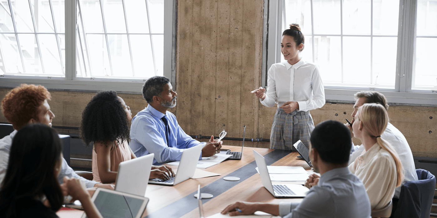 person standing up and speaking to coworkers sitting around a conference room table in the office