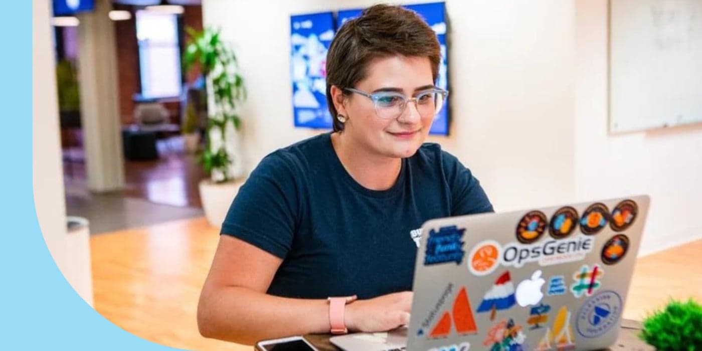 A person sitting in front of an open laptop. They are wearing a blue shirt and glasses. A cell phone is on the table next to their left hand.