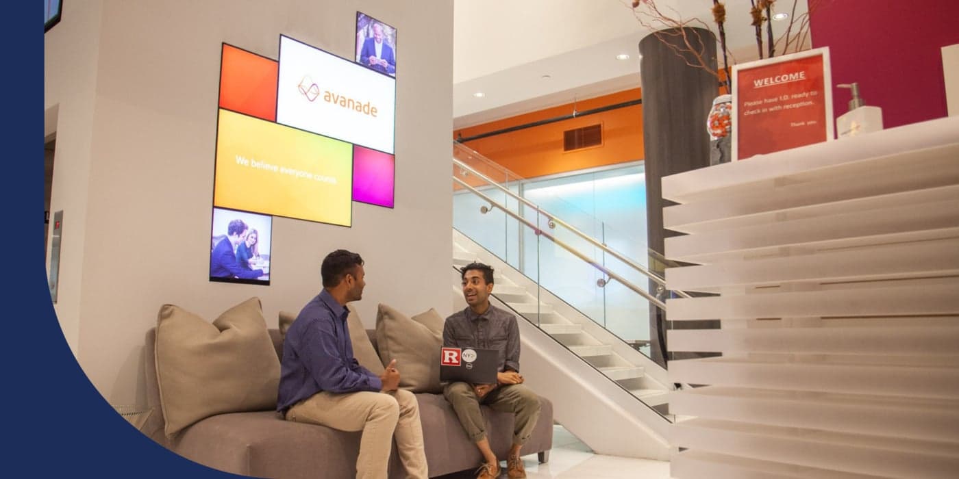 Two people sitting on a light brown couch in an office meeting area. Above them, video screens can be seen. To their right, there is a set of stairs. To the far right, there is a reception desk.