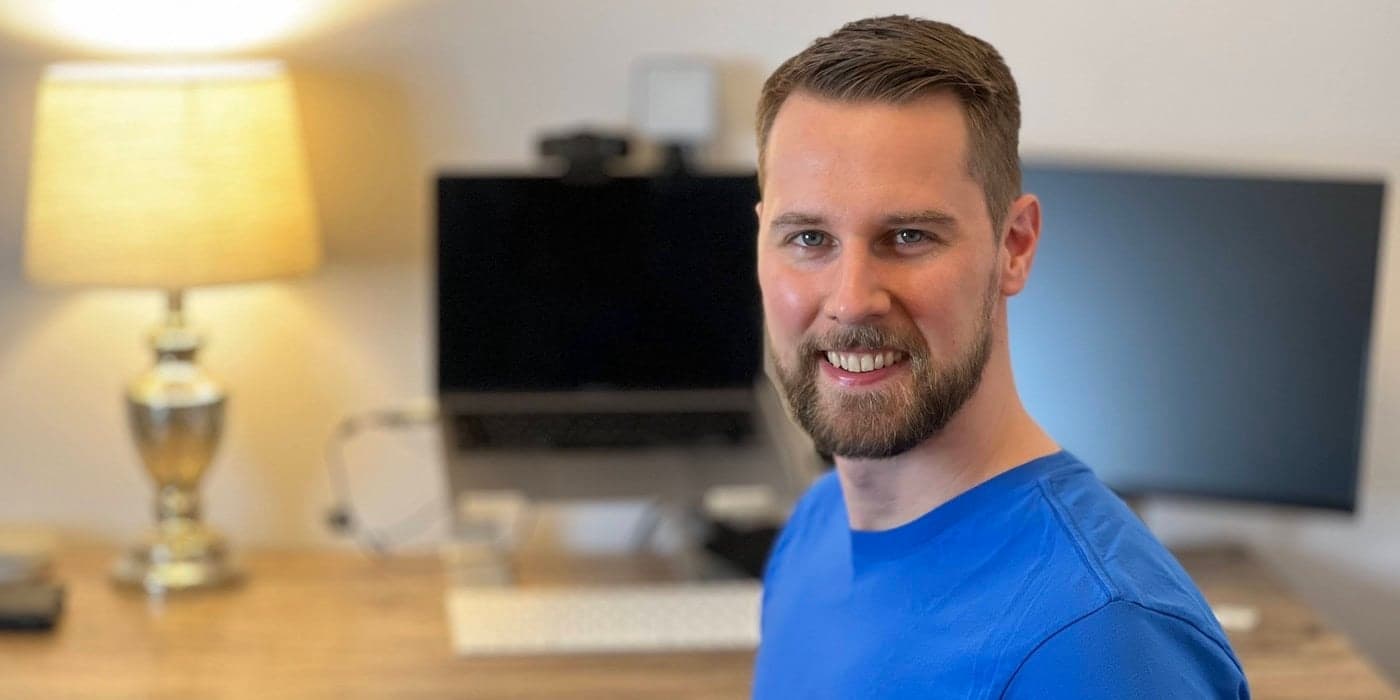 A person with a blue shirt smiling in front of a desk and laptop work-from-home setup.