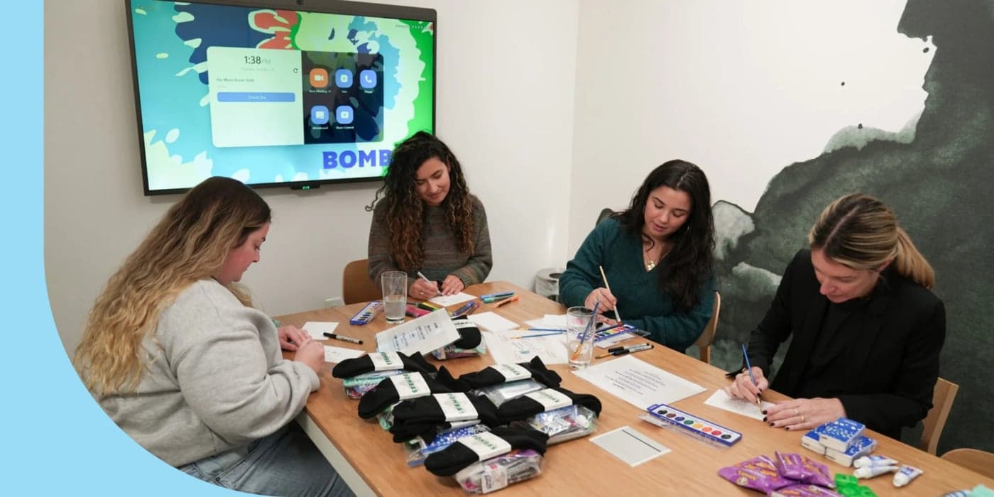 Four people gathered in an office and sat around a table. The person on the far left is wearing a sweatshirt and jeans; packets of black socks are piled in front of them. The other three people are painting pieces of paper using paintbrushes and watercolors. Behind them, a digital screen displays the time.