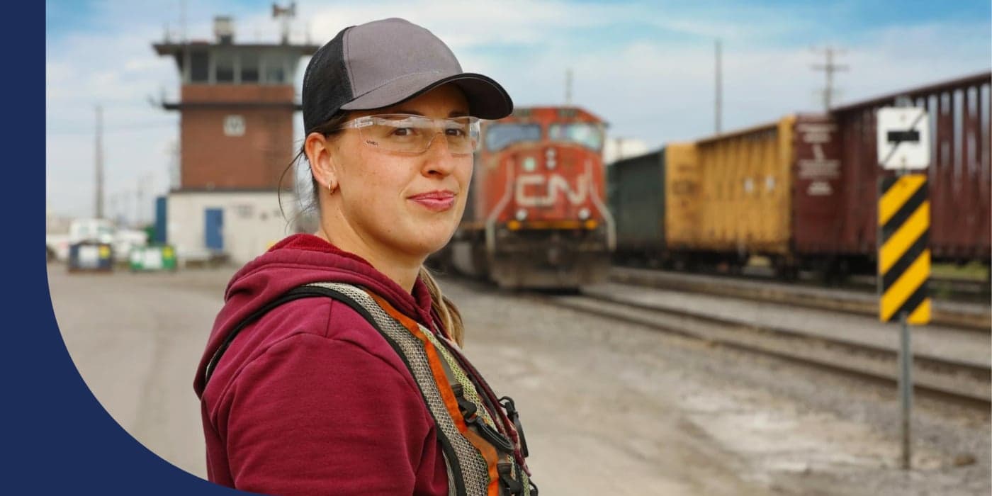 A person standing in a train yard. They are wearing safety glasses, a baseball cap, a red sweatshirt, and a safety vest.