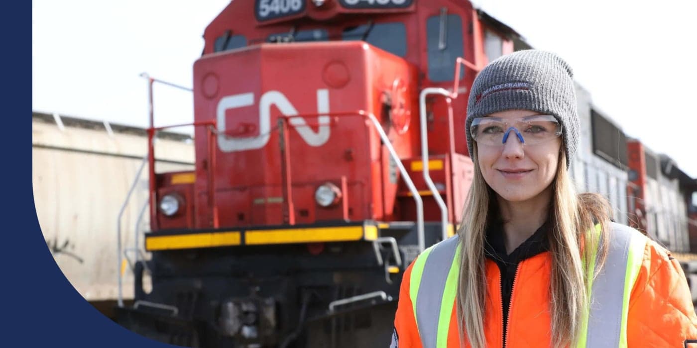 A person dressed in a beanie, safety glasses, and an orange jacket stranding in front of a train. They have long hair.