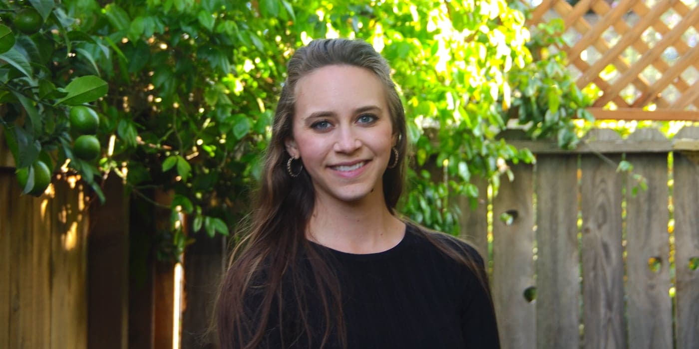 person with long hair standing in front of a tree and picket fence