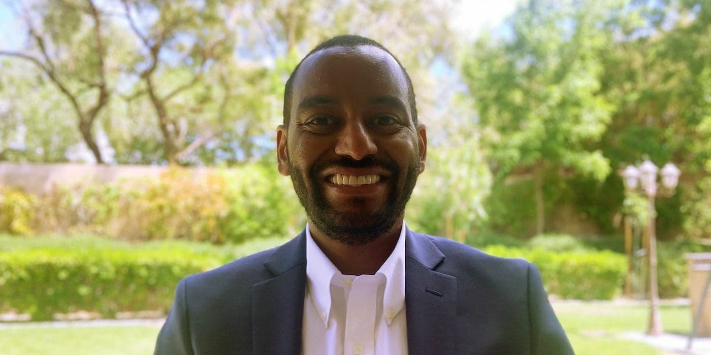 person smiling and standing in front of green bushes and trees