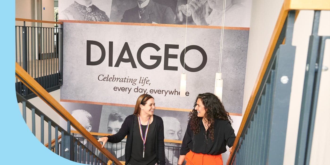 Two smiling people walking up a flight of stairs. The person on the left wears a black shirt, a black sweater, and floral pants. The person on the right wears a black shirt and orange pants. A large banner with the words “DIAGEO” and “Celebrating life, every day, everywhere” is visible behind them.