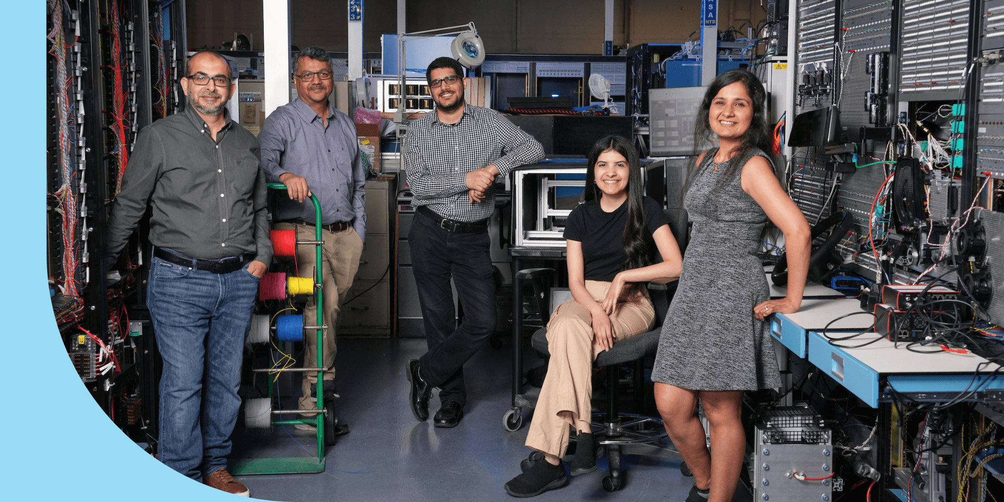 A group of people dressed in business casual attire and standing in an office area. There are computers behind them.