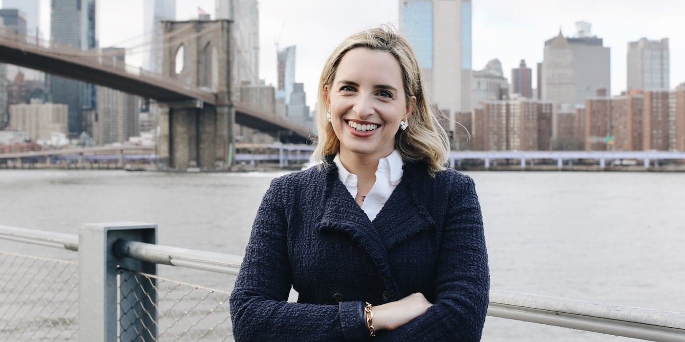 person with blonde hair smiling in front of the Brooklyn Bridge