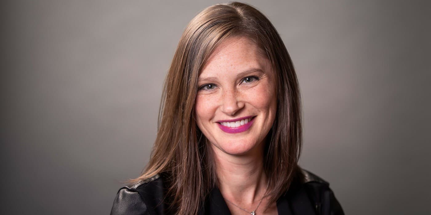 person with straight brown hair smiling against a grey backdrop