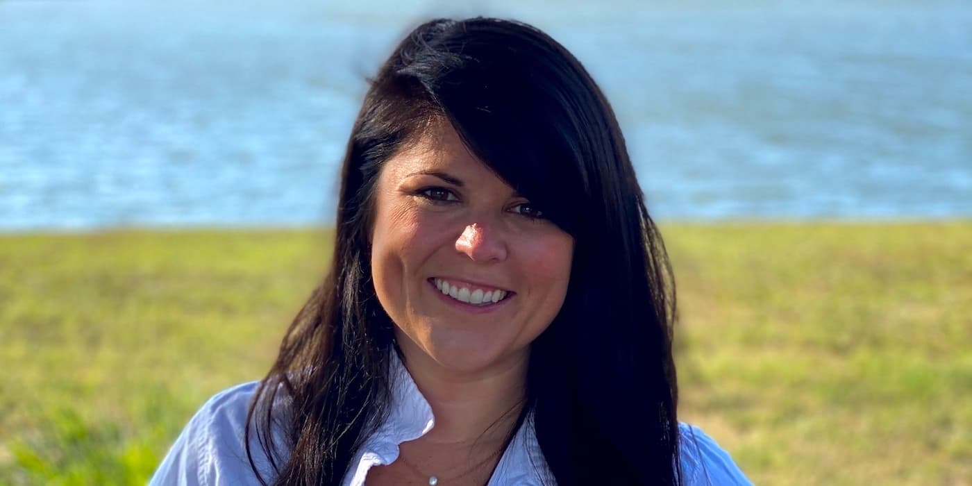 person with long brown hair standing on grass in front of a body of water