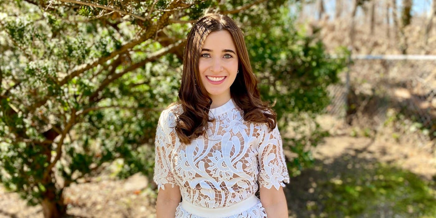 person with brown hair in a white lace dress standing in front of a tree