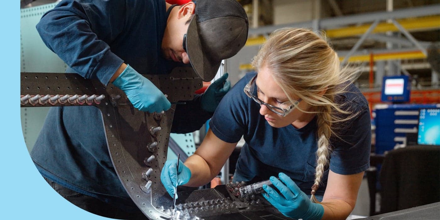 Two people wearing safety glasses and blue gloves while working on a mechanical part. The person on the left is wearing a baseball hat and a long-sleeve shirt; their head is tilted downward. The person on the right has long blonde hair and wears a short-sleeve shirt.