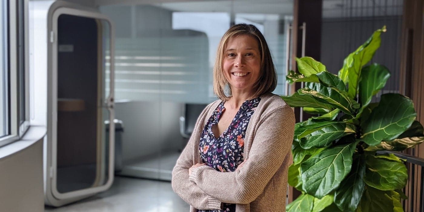 a person with shoulder-length hair smiling with arms crossed in front of a potted plant in an office