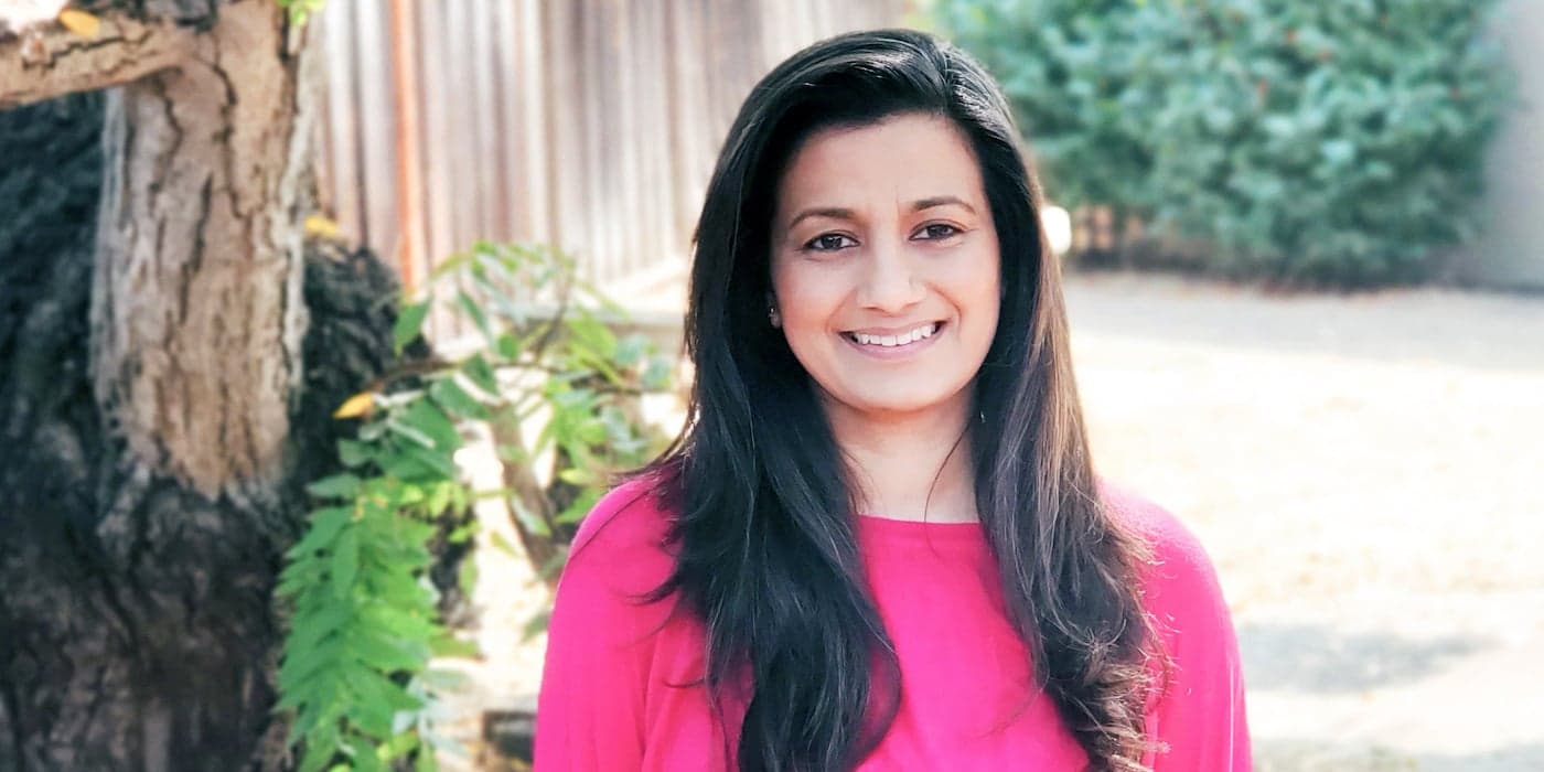person with long brown hair and a pink shirt standing and smiling outside