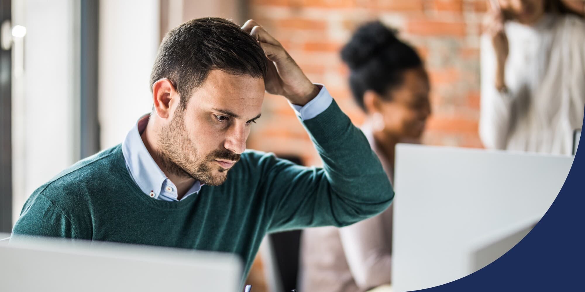 person sitting in an office in front of a computer scratching their head and thinking with a couple of coworkers visible in the background