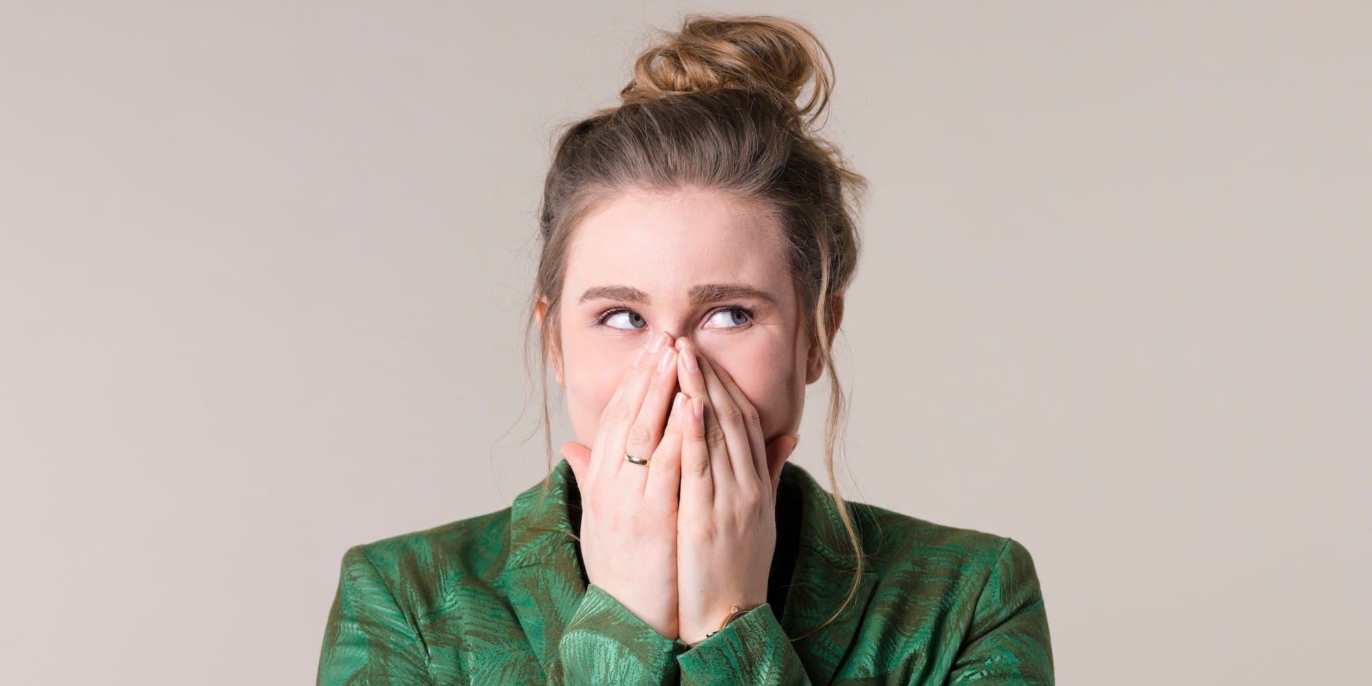 person holding hands over their face and looking off to the side like "oops!" wearing a green top against a beige background
