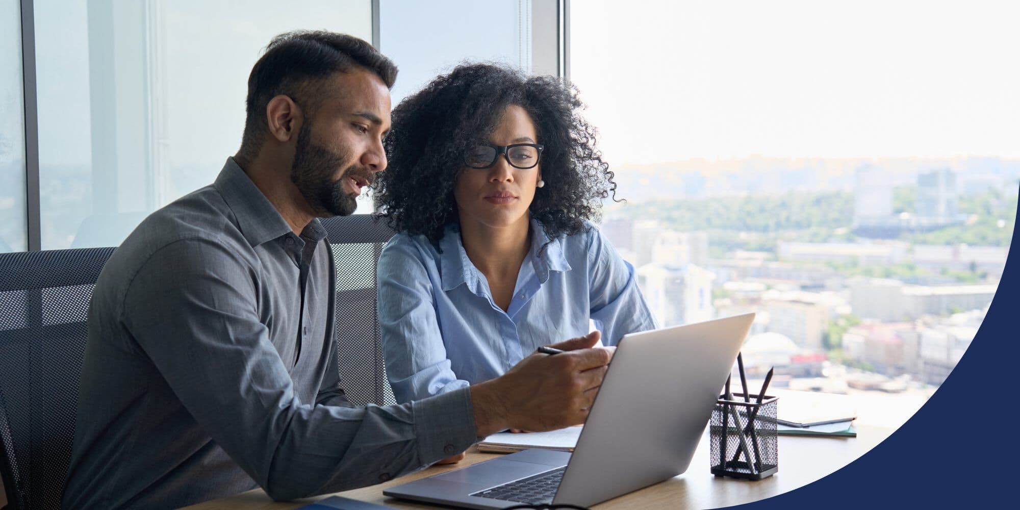 two colleagues of color sitting at a table in an office looking at an open laptop