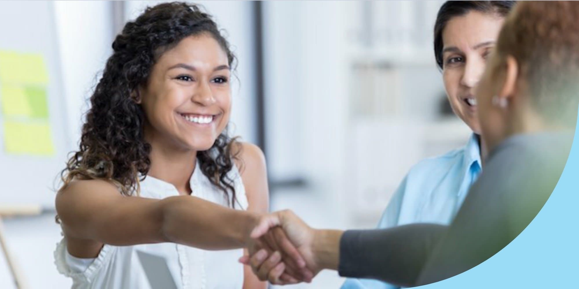 person sitting down facing the camera in a job interview shaking hands with interviewer only partially visible in the frame and one other person visible nearby