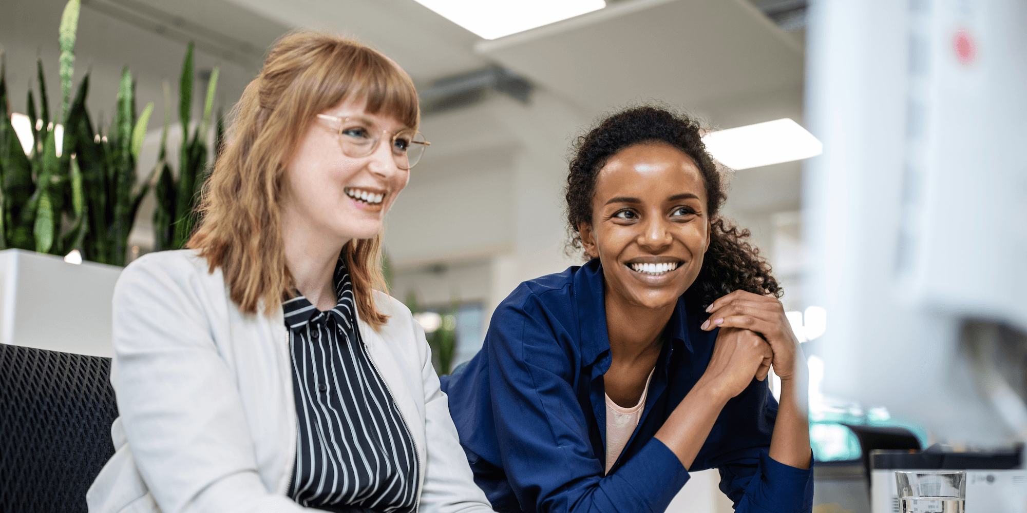 two coworkers in an office looking at a computer screen and smiling, one sitting and one leaning on the desk with their elbows