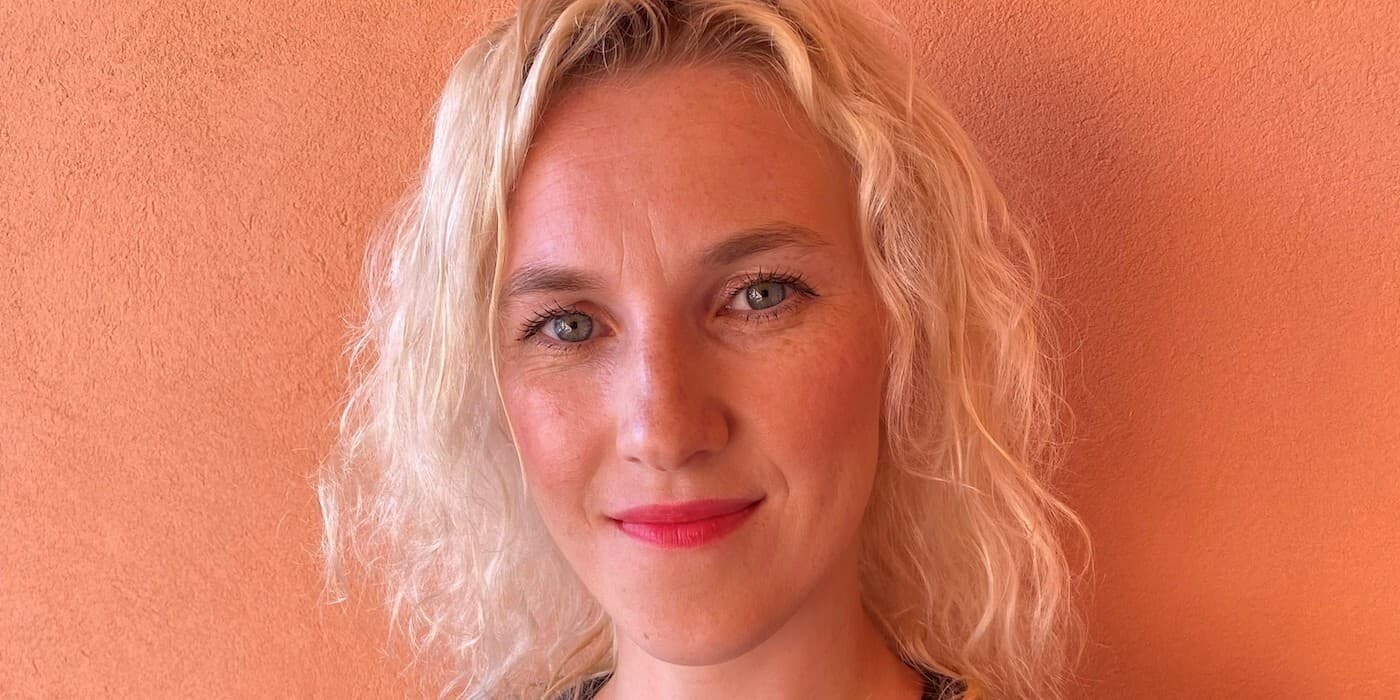 person with short blond wavy hair and blue eyes in front of an orange wall