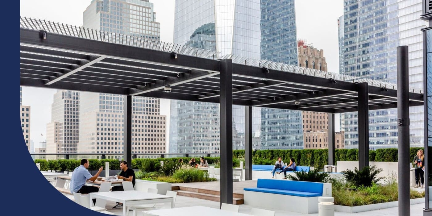 A rooftop area with benches, tables, and chairs. Various pairs of people are sitting at the tables and socializing. Tall office buildings and skyscrapers can be seen in the background.