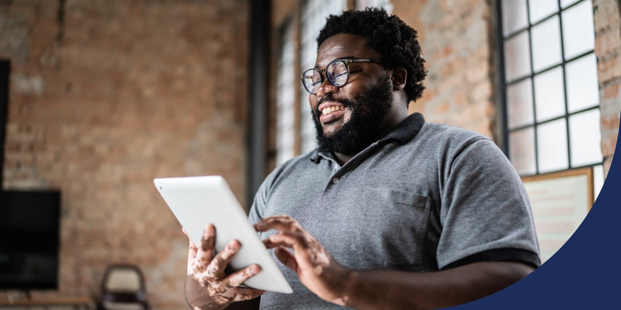 person standing and smiling as they hold and work on a tablet; brick walls and windows visible in the background