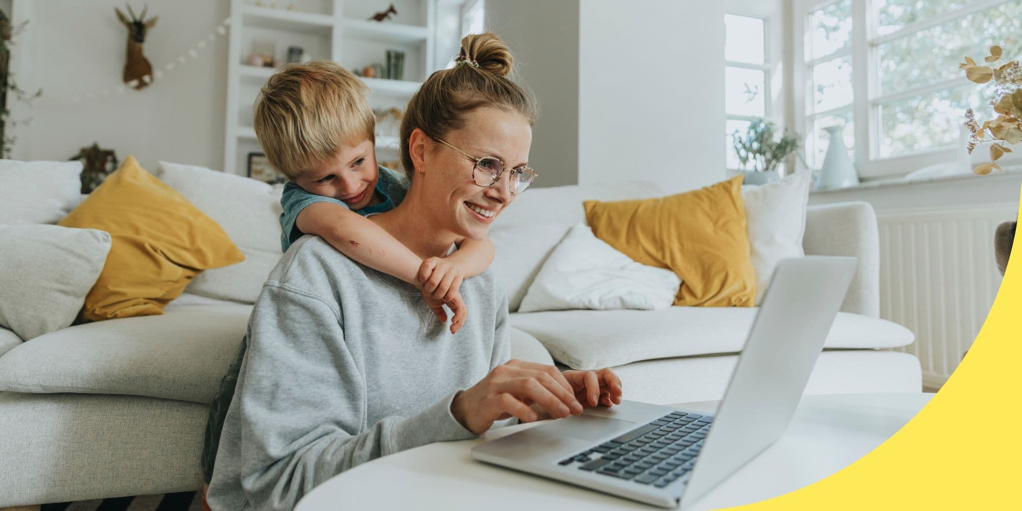 person sitting on the floor between a couch and a coffee table with a child standing behind them, both looking at a laptop on the coffee table