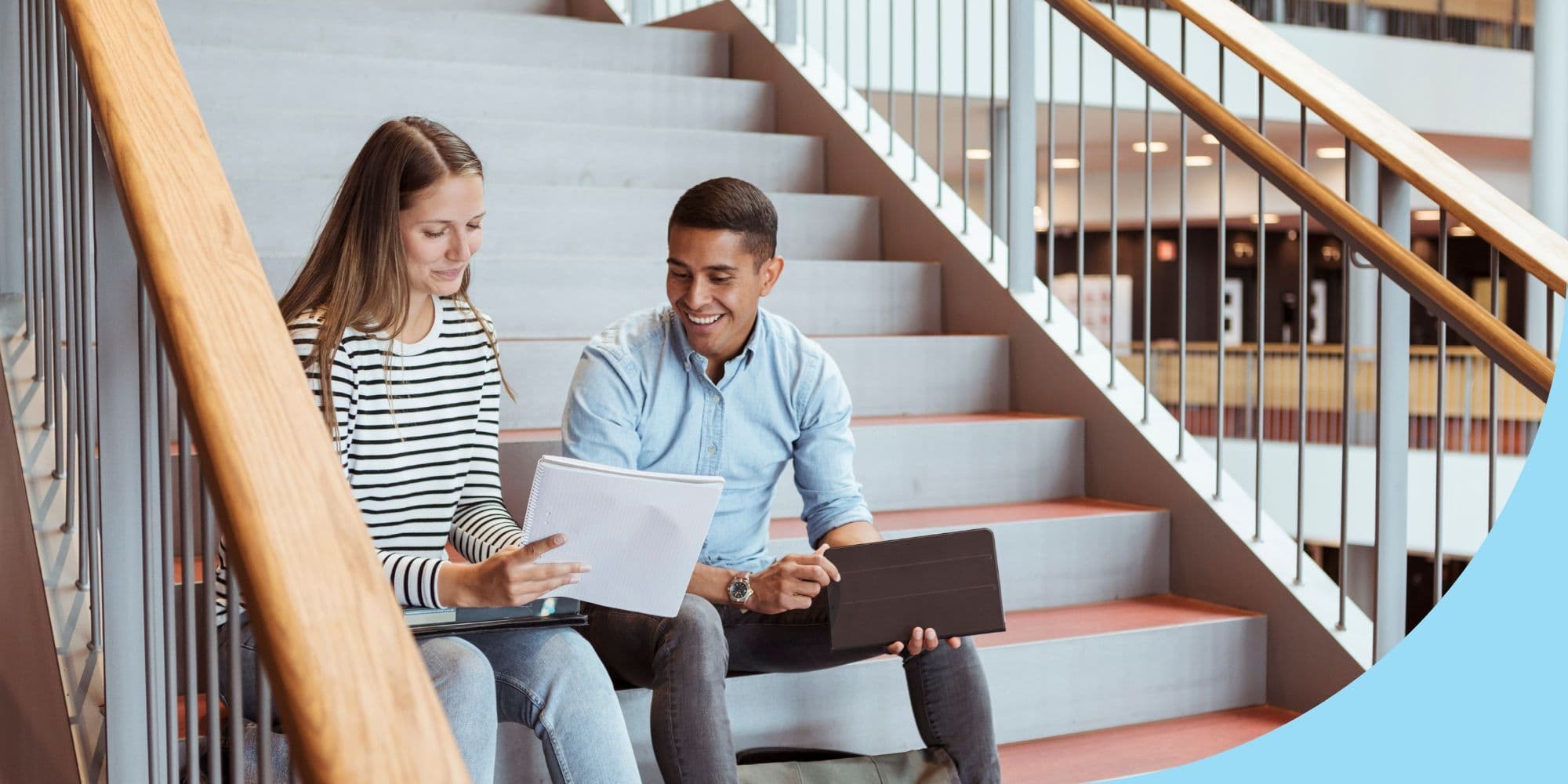 two college students sitting on the stairs talking and looking at a notebook one of them is holding up