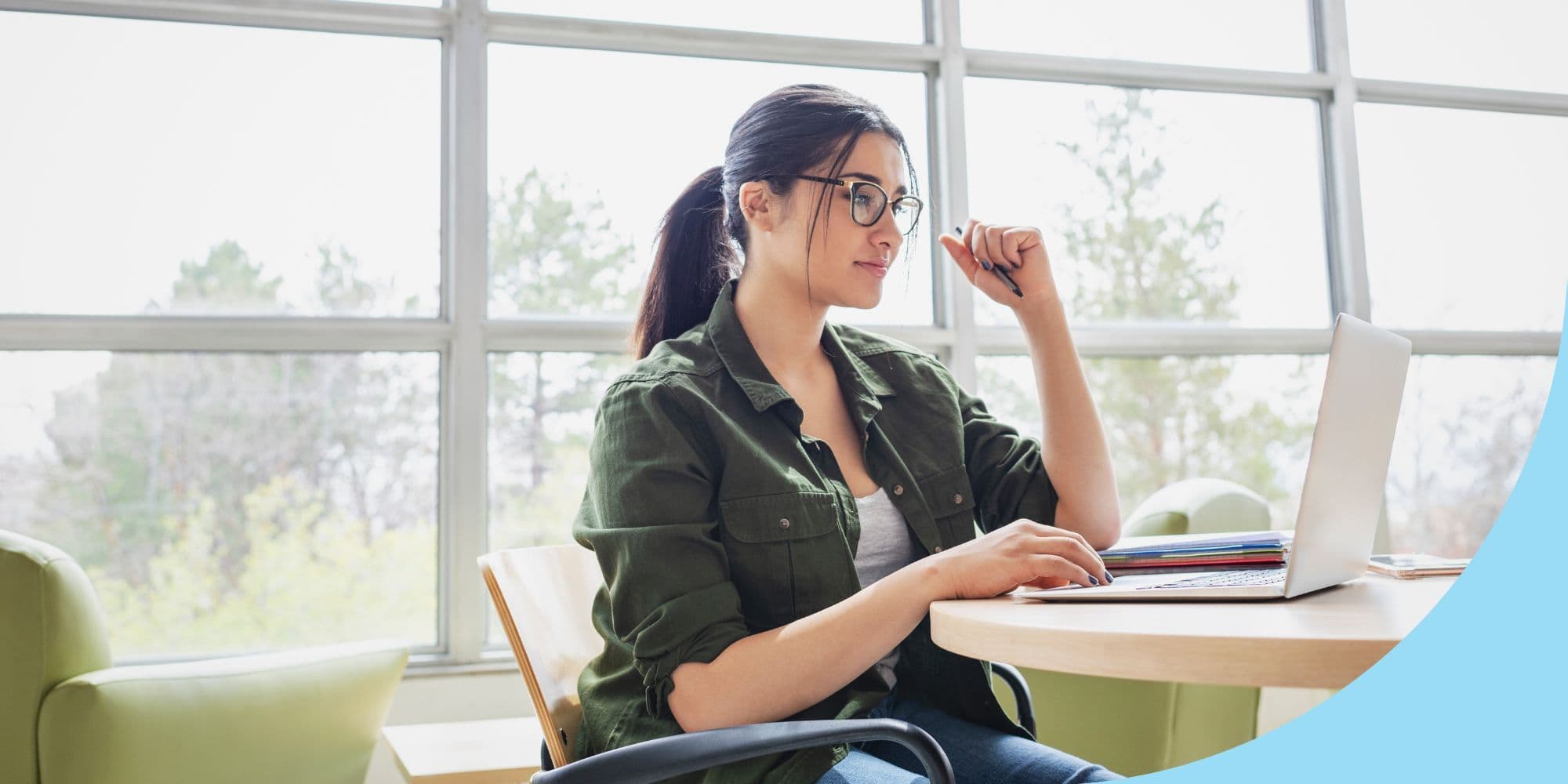 college student sitting at a table looking at a laptop in front of a wall of windows