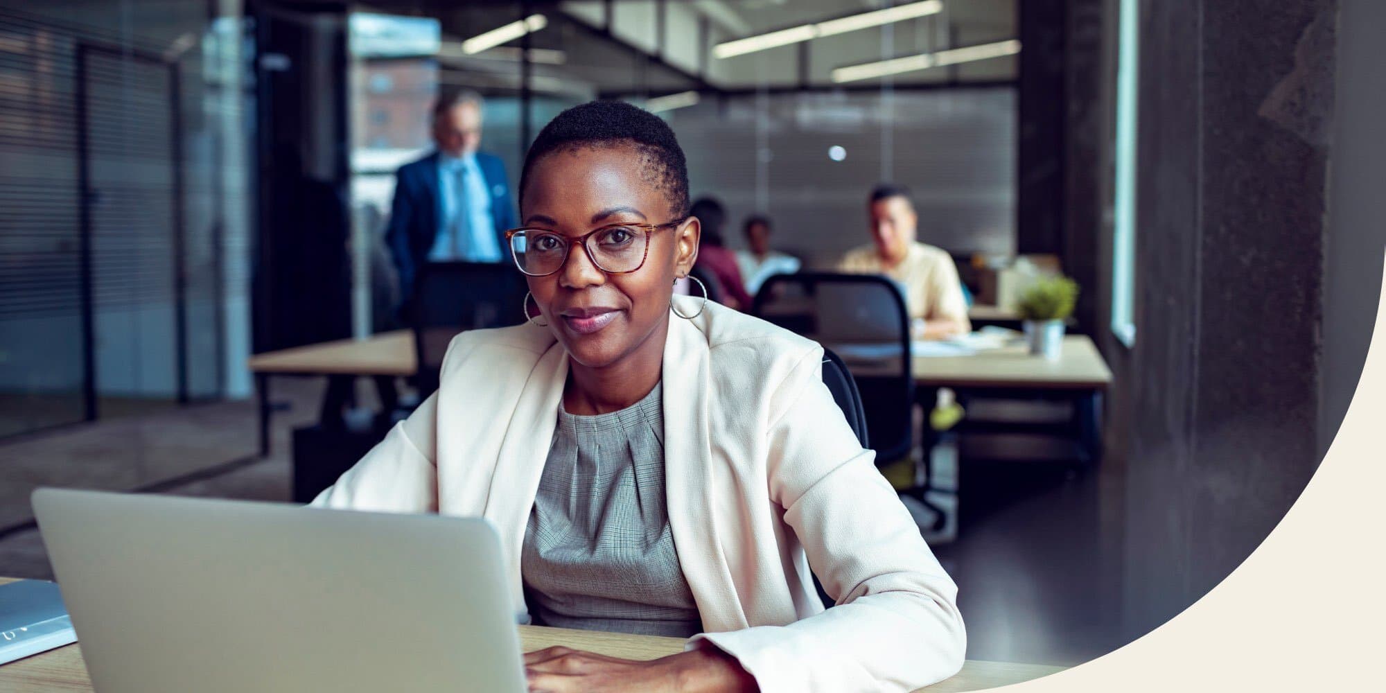 Black woman sitting at a table in an office looking at a laptop with a few colleagues visible in the background